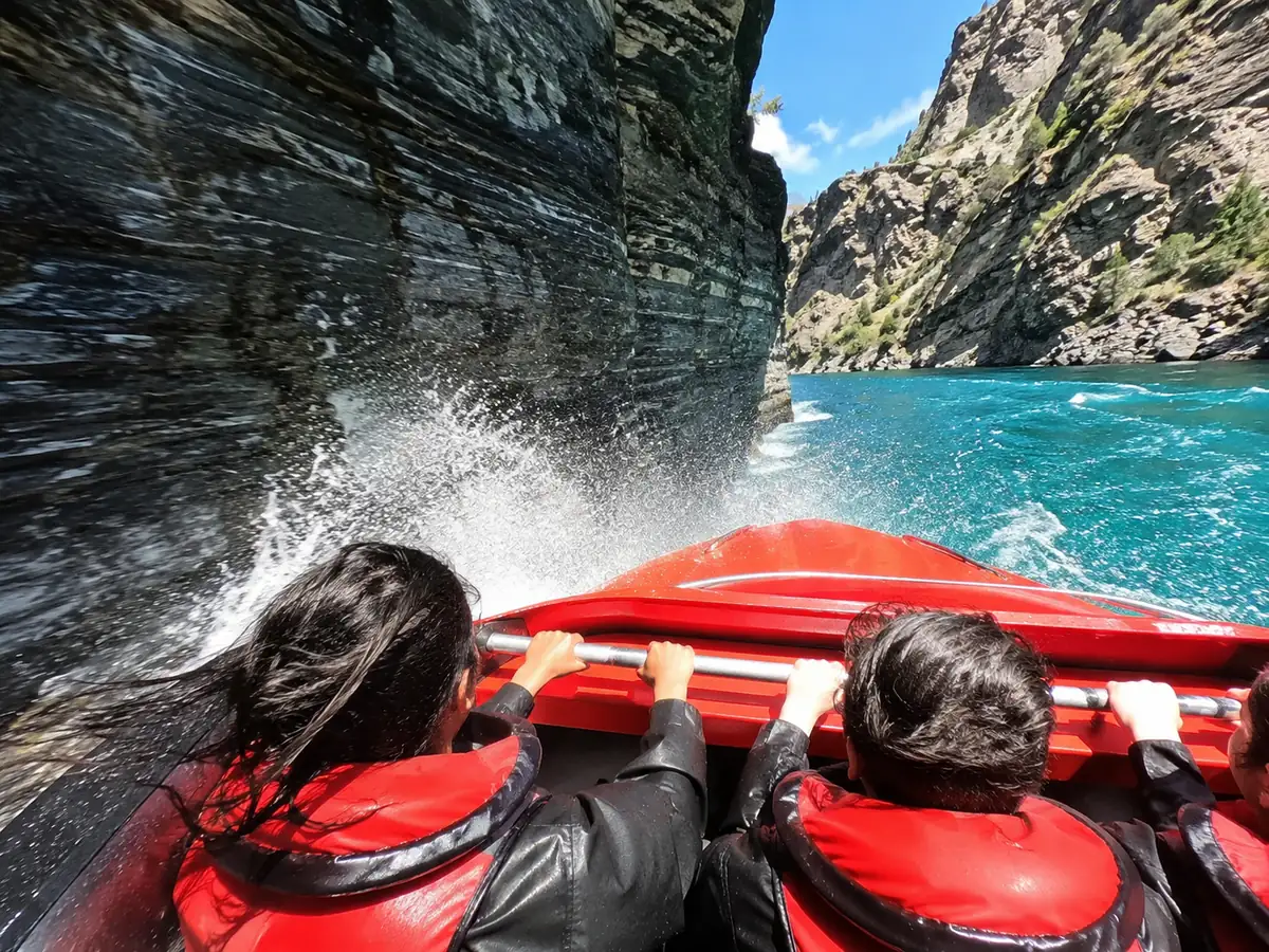 Des passagers agrippés à la rampe tandis qu'un jet boat rouge frôle une paroi de schiste